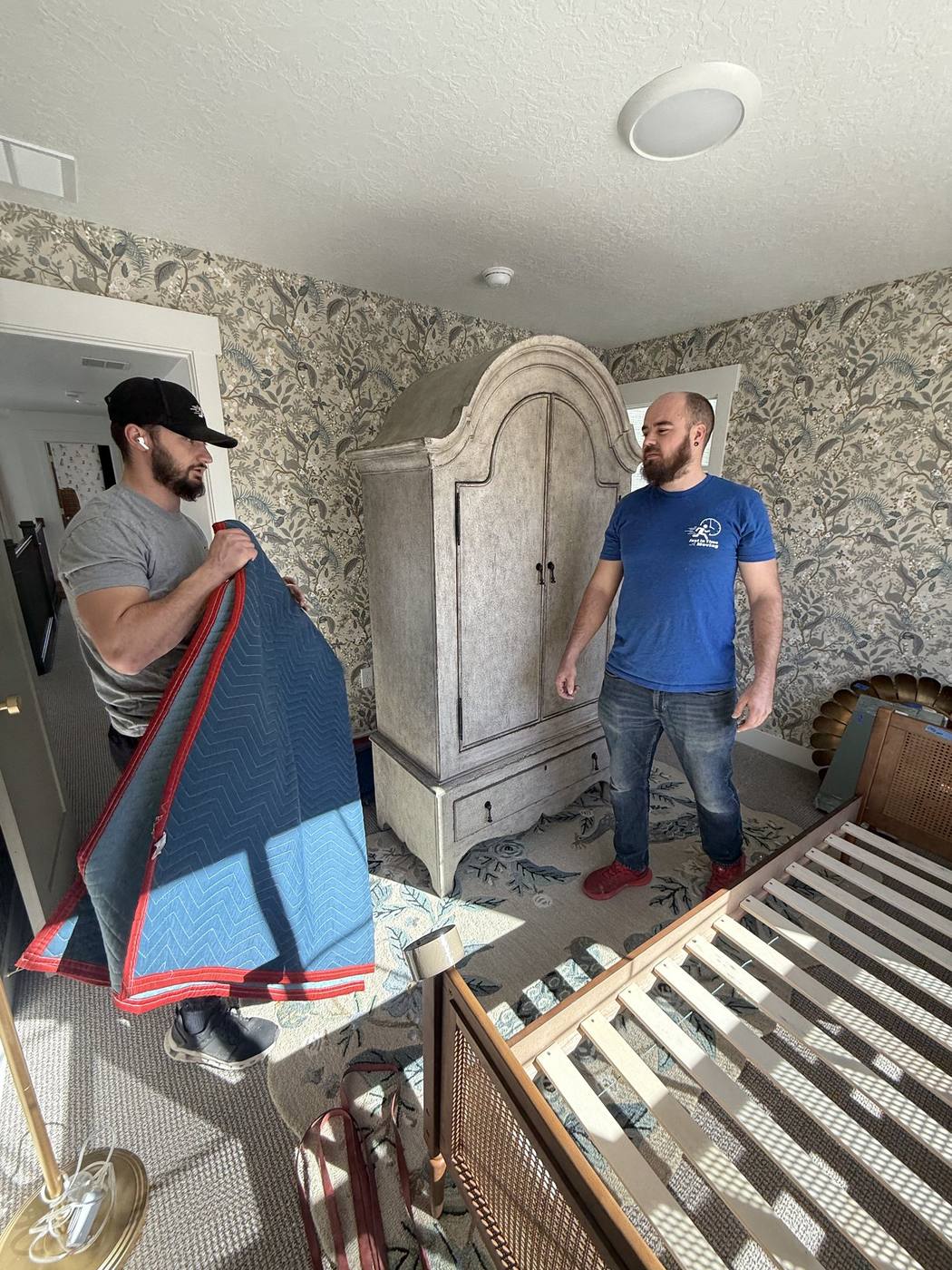 Two Just-In Time movers in branded shirts carefully manoeuvring a tall armoire out of a Northern Utah bedroom